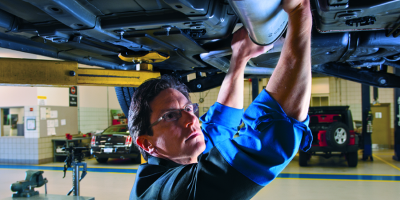 Automotive service technician working underneath a raised vehicle in a service bay, performing maintenance